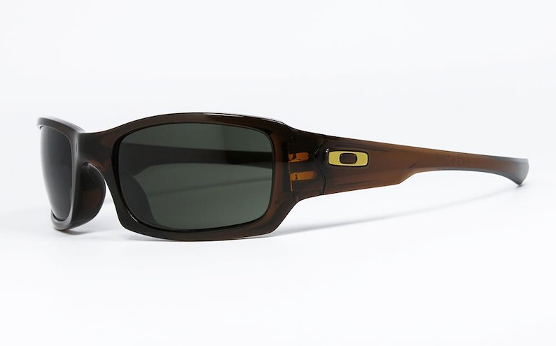 May include: Brown sunglasses with dark lenses. The sunglasses have a rectangular shape with a curved frame. A gold-coloured logo is visible on the temple. The sunglasses are designed for outdoor use.