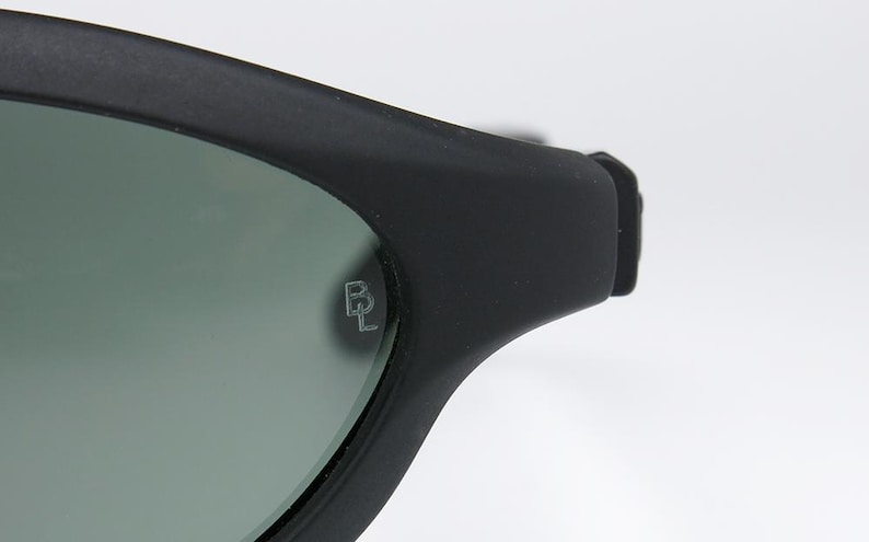 Pode incluir: Close-up de &oacute;culos de sol pretos com acabamento fosco. As lentes s&atilde;o de cor verde suave. O log&oacute;tipo da marca, um "BL" estilizado, est&aacute; gravado na lente. Os &oacute;culos de sol est&atilde;o sobre um fundo branco.
