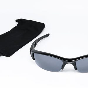 K&ouml;nnte beinhalten: Schwarze Sonnenbrille mit grauen Gl&auml;sern und einer schwarzen Tragetasche. Die Sonnenbrille hat ein schlankes, modernes Design mit einem geschwungenen Rahmen. Die Tasche ist ein weiches, schwarzes Etui.