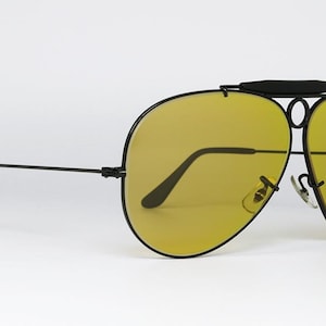 Puede incluir: Gafas de sol de aviador negras con lentes amarillas. Las gafas de sol tienen un doble puente y una barra superior. Las monturas son negras y las lentes son de color amarillo brillante. Las gafas de sol est&aacute;n sobre un fondo blanco.