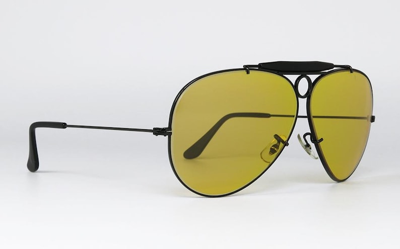 Puede incluir: Gafas de sol de aviador negras con lentes amarillas. Las gafas de sol tienen un doble puente y una barra superior. Las monturas son negras y las lentes son de color amarillo brillante. Las gafas de sol est&aacute;n sobre un fondo blanco.