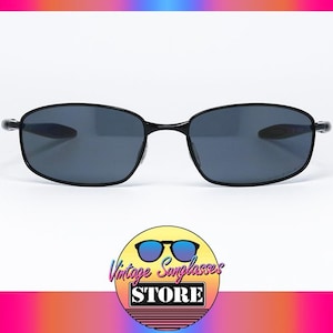 Puede incluir: Gafas de sol negras con lentes de color gris oscuro. Las gafas tienen forma rectangular y una montura fina. La imagen incluye un logotipo en la parte inferior que dice "Vintage Sunglasses Store". El fondo es blanco con un degradado de color.