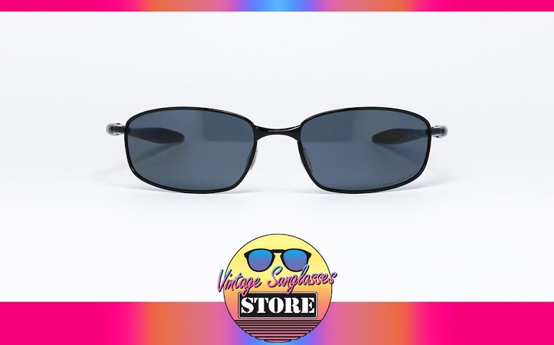 Puede incluir: Gafas de sol negras con lentes de color gris oscuro. Las gafas tienen forma rectangular y una montura fina. La imagen incluye un logotipo en la parte inferior que dice "Vintage Sunglasses Store". El fondo es blanco con un degradado de color.