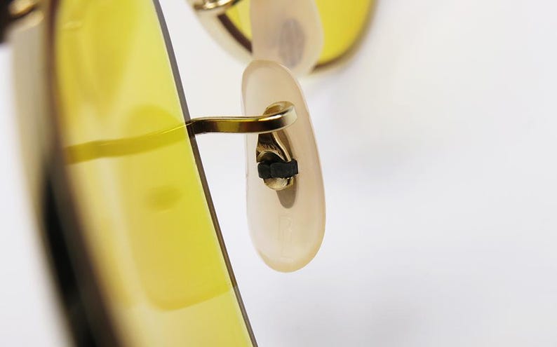 K&ouml;nnte beinhalten: Nahaufnahme des goldfarbenen Metall-Scharniers und des B&uuml;gelendes einer Sonnenbrille mit gelber T&ouml;nung.