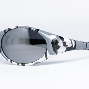 May include: Sport sunglasses with a gray and white camouflage pattern on the frames and temples. The lenses are a dark gray color. The sunglasses have a curved design and a black ear piece.