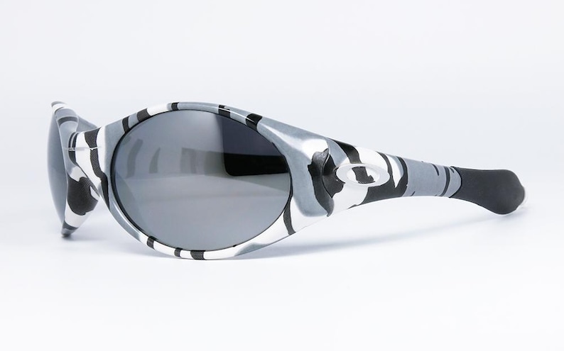 May include: Sport sunglasses with a gray and white camouflage pattern on the frames and temples. The lenses are a dark gray color. The sunglasses have a curved design and a black ear piece.