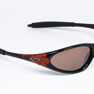 Pode incluir: &Oacute;culos de sol pretos com lentes castanhas. As arma&ccedil;&otilde;es s&atilde;o pretas com um desenho floral em laranja e castanho nas hastes. As hastilhas s&atilde;o castanhas escuras com extremidades pretas. O log&oacute;tipo Oakley &eacute; vis&iacute;vel.