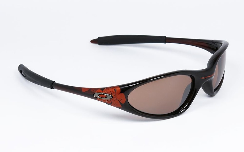 Pode incluir: &Oacute;culos de sol pretos com lentes castanhas. As arma&ccedil;&otilde;es s&atilde;o pretas com um desenho floral em laranja e castanho nas hastes. As hastilhas s&atilde;o castanhas escuras com extremidades pretas. O log&oacute;tipo Oakley &eacute; vis&iacute;vel.