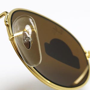 Pu&ograve; includere: Primo piano di occhiali da sole con montatura dorata e lenti marroni. Gli occhiali da sole hanno naselli trasparenti e un design classico. Le lenti sono di forma ovale e la montatura &egrave; dorata.