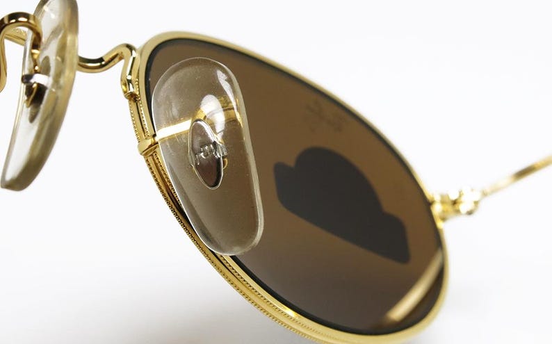 Pu&ograve; includere: Primo piano di occhiali da sole con montatura dorata e lenti marroni. Gli occhiali da sole hanno naselli trasparenti e un design classico. Le lenti sono di forma ovale e la montatura &egrave; dorata.