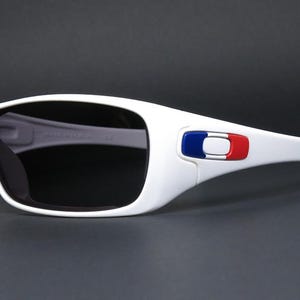Op de afbeelding: Witte zonnebril met donkere glazen. De monturen hebben een blauw, wit en rood embleem op de slapen. De zonnebril heeft een sportief design en staat tegen een donkere achtergrond.