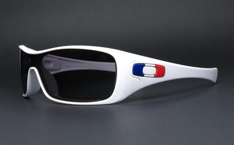 Op de afbeelding: Witte zonnebril met donkere glazen. De monturen hebben een blauw, wit en rood embleem op de slapen. De zonnebril heeft een sportief design en staat tegen een donkere achtergrond.