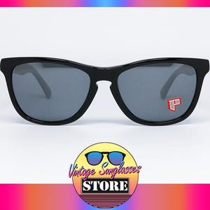 May include: Black sunglasses with gray lenses. The sunglasses have a rectangular frame and a small red and white logo on the right lens. The image also includes a logo for the Vintage Sunglasses Store.
