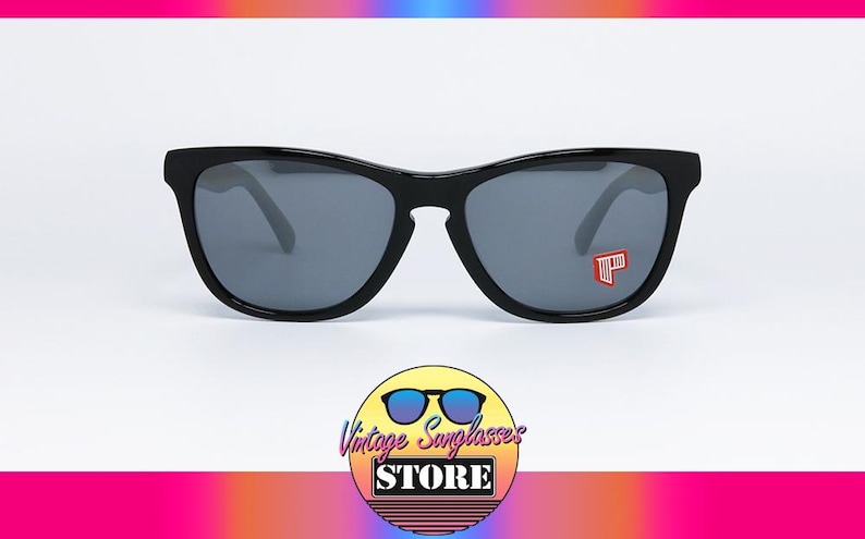 May include: Black sunglasses with gray lenses. The sunglasses have a rectangular frame and a small red and white logo on the right lens. The image also includes a logo for the Vintage Sunglasses Store.