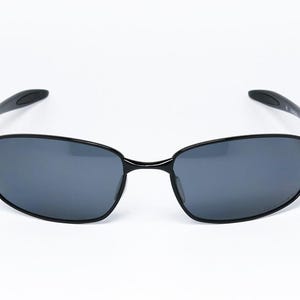 Puede incluir: Gafas de sol negras con montura rectangular y lentes gris oscuro. Las patillas son curvas y se extienden hasta los terminales. Las gafas de sol est&aacute;n sobre un fondo blanco.