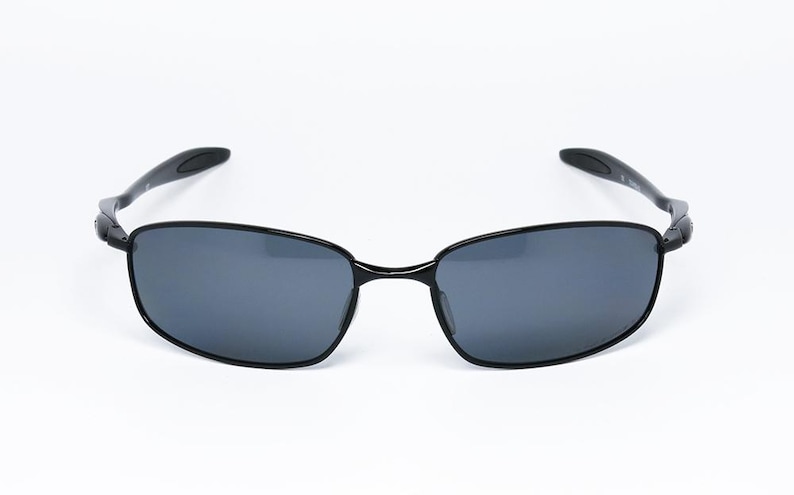 Puede incluir: Gafas de sol negras con montura rectangular y lentes gris oscuro. Las patillas son curvas y se extienden hasta los terminales. Las gafas de sol est&aacute;n sobre un fondo blanco.