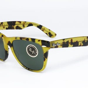 May include: Tortoiseshell sunglasses with dark lenses. The frames are a mix of yellow and brown, with the Ray-Ban logo visible on the arm. The lenses are a dark green color.