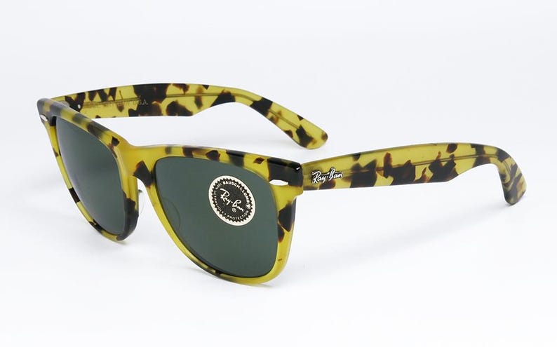 May include: Tortoiseshell sunglasses with dark lenses. The frames are a mix of yellow and brown, with the Ray-Ban logo visible on the arm. The lenses are a dark green color.