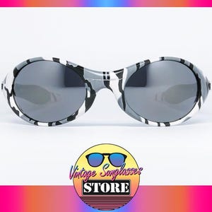 May include: Oval-shaped sunglasses with a black, white, and gray camouflage pattern on the frames. The lenses are a dark gray color. The sunglasses are displayed on a white background. The logo for Vintage Sunglasses Store is in the bottom center.