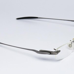 May include: A pair of rimless eyeglasses with a silver frame and black temple tips. The glasses have a flexible hinge and a clear lens.