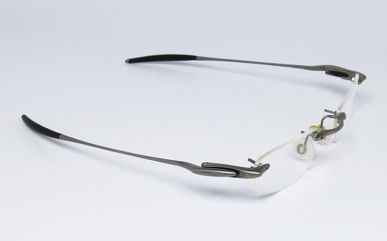 May include: A pair of rimless eyeglasses with a silver frame and black temple tips. The glasses have a flexible hinge and a clear lens.