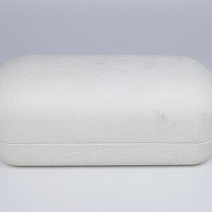 May include: A white, rectangular eyeglasses case with rounded edges. The case has a textured, embossed floral pattern across its surface. The case is closed and sits on a white background.