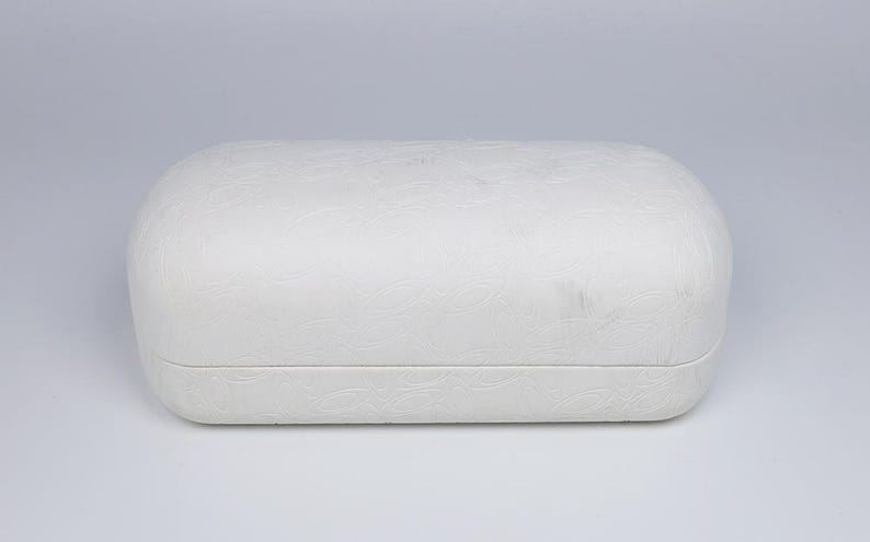 May include: A white, rectangular eyeglasses case with rounded edges. The case has a textured, embossed floral pattern across its surface. The case is closed and sits on a white background.