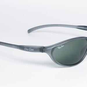 May include: Gray Ray-Ban sunglasses with dark green lenses. The sunglasses have a curved design and the Ray-Ban logo is visible on the temples and lens. The frame is a matte gray color.