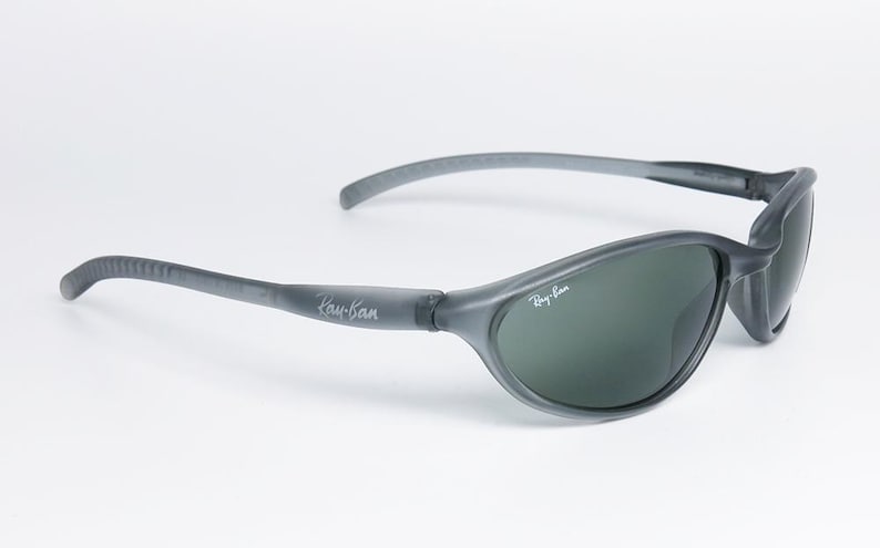 May include: Gray Ray-Ban sunglasses with dark green lenses. The sunglasses have a curved design and the Ray-Ban logo is visible on the temples and lens. The frame is a matte gray color.