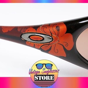 Pode incluir: Close-up de &oacute;culos de sol com arma&ccedil;&atilde;o preta e lentes castanhas. As hastes apresentam um design floral laranja e vermelho, com um log&oacute;tipo prateado. &Oacute;culos de sol da Vintage Sunglasses Store.
