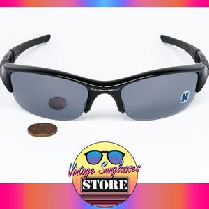 K&ouml;nnte beinhalten: Schwarze Sonnenbrille mit grauen Gl&auml;sern. Die Sonnenbrille hat ein halb rahmenloses Design und einen geschwungenen Rahmen. Neben der Sonnenbrille liegt ein Cent-St&uuml;ck. Das Bild enth&auml;lt auch ein Logo f&uuml;r einen Vintage-Sonnenbrillenladen.