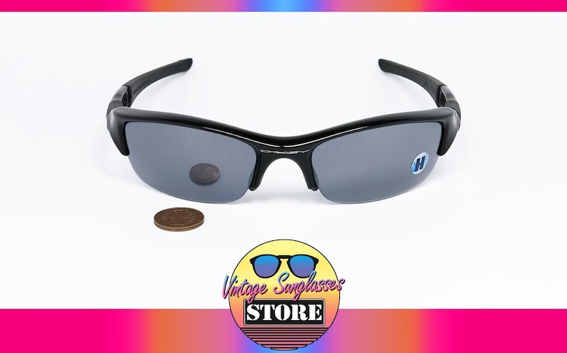 K&ouml;nnte beinhalten: Schwarze Sonnenbrille mit grauen Gl&auml;sern. Die Sonnenbrille hat ein halb rahmenloses Design und einen geschwungenen Rahmen. Neben der Sonnenbrille liegt ein Cent-St&uuml;ck. Das Bild enth&auml;lt auch ein Logo f&uuml;r einen Vintage-Sonnenbrillenladen.