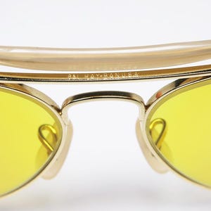 K&ouml;nnte beinhalten: Goldfarbene Aviator-Sonnenbrille mit gelben Gl&auml;sern. Die Sonnenbrille hat einen Metallrahmen und eine Br&uuml;cke mit der Aufschrift "Ray-Ban USA".