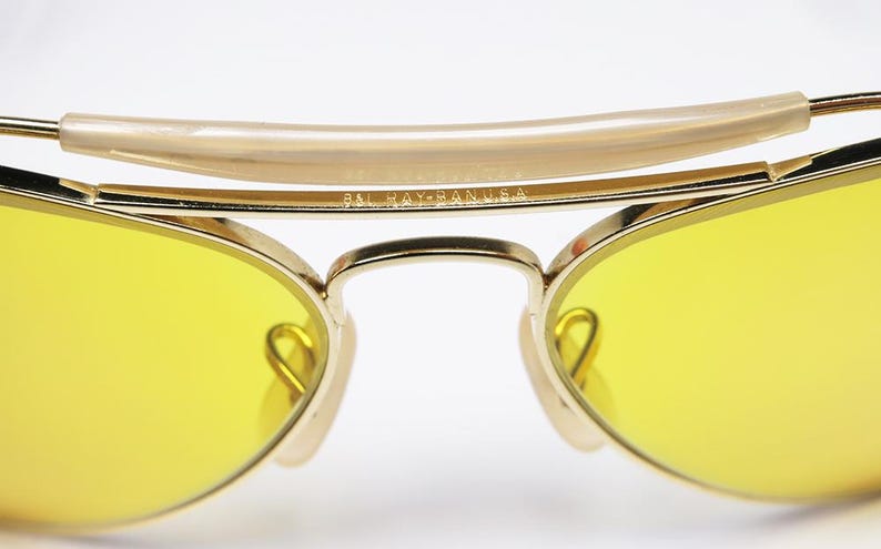K&ouml;nnte beinhalten: Goldfarbene Aviator-Sonnenbrille mit gelben Gl&auml;sern. Die Sonnenbrille hat einen Metallrahmen und eine Br&uuml;cke mit der Aufschrift "Ray-Ban USA".