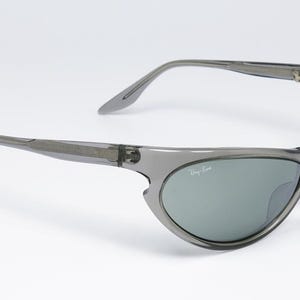 May include: Gray sunglasses with a wraparound design. The lenses are a muted green color, and the Ray-Ban logo is visible on the lens. The frame is a translucent gray plastic.