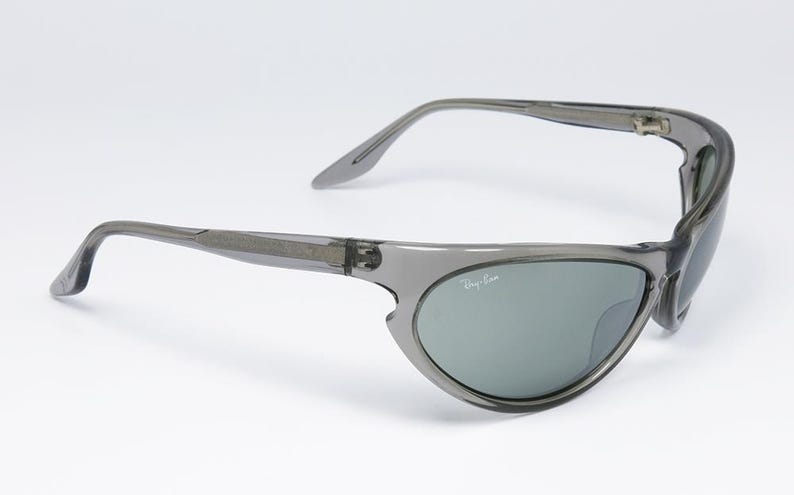 May include: Gray sunglasses with a wraparound design. The lenses are a muted green color, and the Ray-Ban logo is visible on the lens. The frame is a translucent gray plastic.
