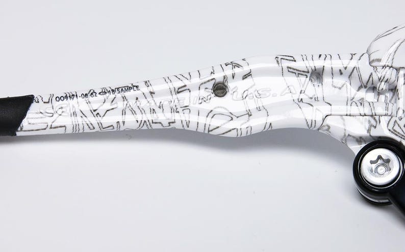 May include: White and black bicycle brake lever with a graphic design. The lever has a black grip and a black bolt. The text "004171-06 62" and "SAMPLE" is printed on the lever.