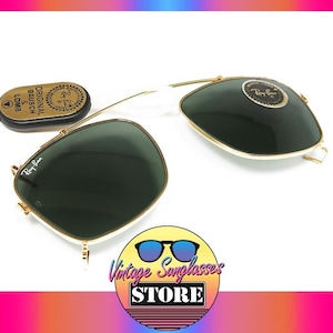 May include: Ray-Ban sunglasses with dark green lenses and gold frames, along with a small, gold-colored case. The sunglasses are displayed on a white surface. The logo of the Vintage Sunglasses Store is in the bottom right corner.
