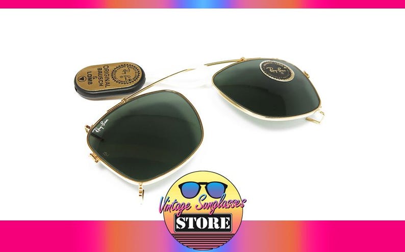 May include: Ray-Ban sunglasses with dark green lenses and gold frames, along with a small, gold-colored case. The sunglasses are displayed on a white surface. The logo of the Vintage Sunglasses Store is in the bottom right corner.