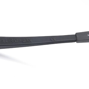 Pode incluir: Close-up de uma haste de &oacute;culos de sol Ray-Ban preta. O texto "B & L RAY-BAN" est&aacute; em relevo na haste. O texto "MADE IN IRELAND" tamb&eacute;m &eacute; vis&iacute;vel. A haste dos &oacute;culos de sol est&aacute; em um fundo branco.