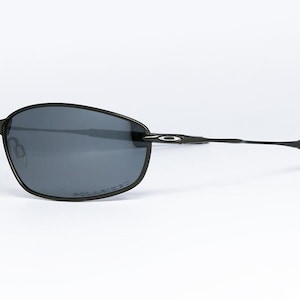 Może przedstawiać: Czarne okulary przeciwsłoneczne o eleganckim, nowoczesnym designie. Soczewki są w kolorze ciemnoszarym, a oprawki z czarnego metalu. Zaśnki są cienkie i lekko zakrzywione na końcach. Okulary są na białym tle.