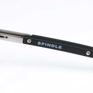 May include: Close-up of eyeglasses with a black frame and silver hinges. The temple arm is black with the word "SPINDLE" printed in white. The glasses are set against a white background.