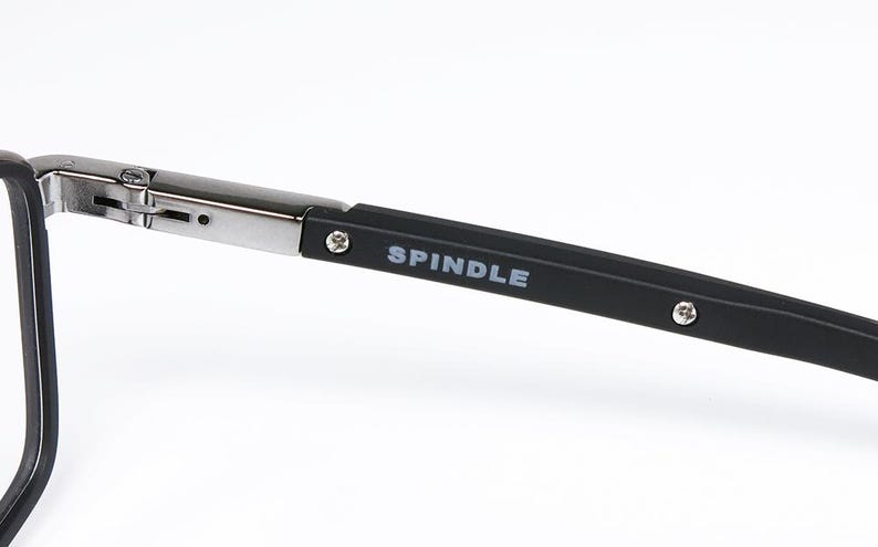 May include: Close-up of eyeglasses with a black frame and silver hinges. The temple arm is black with the word "SPINDLE" printed in white. The glasses are set against a white background.