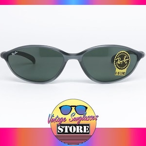 May include: Dark gray Ray-Ban sunglasses with dark lenses. The sunglasses have a curved frame and a small Ray-Ban logo on the temple. A gold sticker with the text "G-15 LENS" is on the right lens. The sunglasses are displayed on a white background.