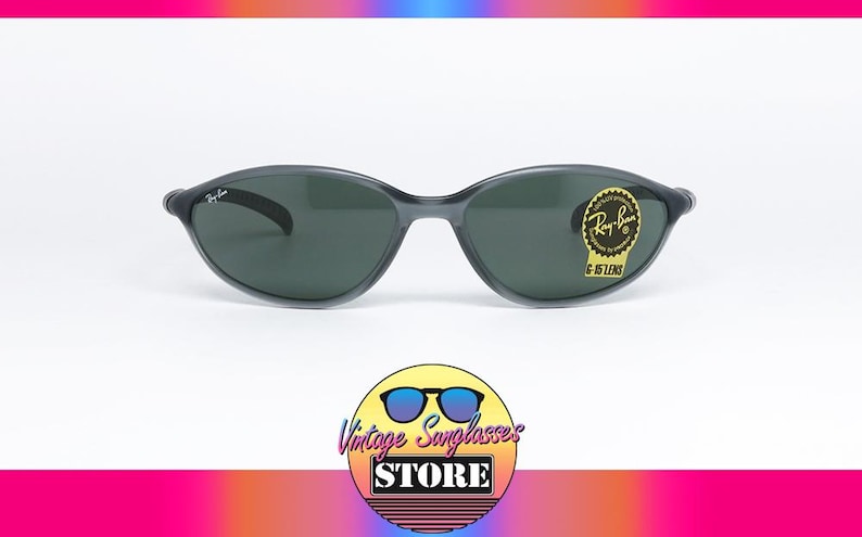 May include: Dark gray Ray-Ban sunglasses with dark lenses. The sunglasses have a curved frame and a small Ray-Ban logo on the temple. A gold sticker with the text "G-15 LENS" is on the right lens. The sunglasses are displayed on a white background.