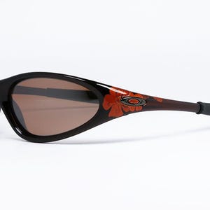 Pode incluir: &Oacute;culos de sol pretos e castanhos com um design floral nas hastes. As lentes s&atilde;o castanhas. Os &oacute;culos t&ecirc;m um estilo desportivo e envolvente.