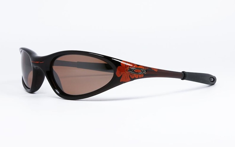 Pode incluir: &Oacute;culos de sol pretos e castanhos com um design floral nas hastes. As lentes s&atilde;o castanhas. Os &oacute;culos t&ecirc;m um estilo desportivo e envolvente.