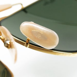 May include: Close-up of gold-rimmed sunglasses with green lenses. The image highlights the nose pads and the frame's details. The lenses are dark green, and the nose pads are a light beige color.