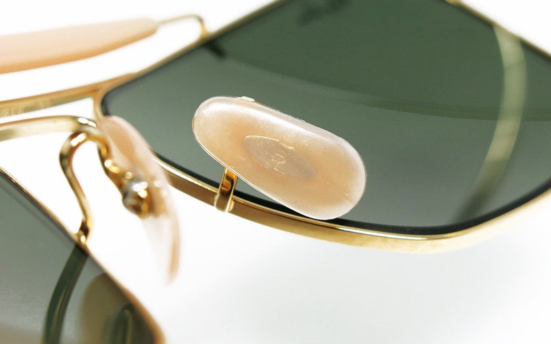 May include: Close-up of gold-rimmed sunglasses with green lenses. The image highlights the nose pads and the frame's details. The lenses are dark green, and the nose pads are a light beige color.