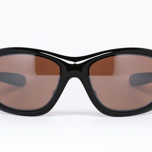 K&ouml;nnte beinhalten: Schwarze Sonnenbrille mit braunen Gl&auml;sern. Die Sonnenbrille hat einen dicken, abgerundeten Rahmen und ein geschwungenes Design. Die Gl&auml;ser sind warmbraun und der Rahmen ist gl&auml;nzend schwarz. Die Sonnenbrille ist vor einem wei&szlig;en Hintergrund platziert.